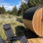 Holzsauna mit offenem Eingang auf Holzterrasse, zwei schwarze Liegestühle im Gras, Hintergrund mit Bäumen.