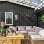 Terrace with seating group, wooden table, and plants against black wood wall under glass roof.