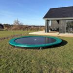 Rundes Trampolin auf grünem Rasen vor einem Holzhaus mit Terrasse.