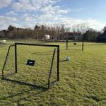 Zwei Fußballtore und ein Ball auf einem grünen Rasenfeld unter blauem Himmel.