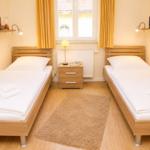 Zwei Einzelbetten mit weißen Laken, Nachttisch und Fenster mit gelben Vorhängen.