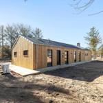 Moderne Holzhütte mit mehreren Eingängen und Fenstern, umgeben von Bäumen und freiem Gelände.