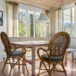 Dining area with wooden table, rattan chairs, and bench. Large windows, refrigerator, and bookshelf.