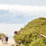 Family walks along dune path to sea promenade. Bicycle and traffic signs are visible.