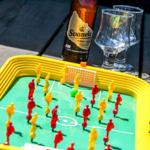 Foosball table with red and yellow figures, bottle and two glasses on wooden table.