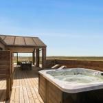 Terrasse mit Whirlpool, Grill und Sitzgelegenheiten. Blick auf offenes Feld unter blauem Himmel.