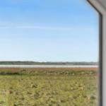 Aus dem Fenster blickt man auf eine Wiese mit Gras und gelben Blumen, dahinter ein Fluss und eine grüne Landschaft.