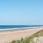 Strand mit Sanddünen und Meer unter blauem Himmel. Ein einzelner Mensch am Ufer.