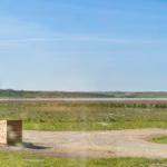 Draußen liegt ein Holzobjekt neben einem unbefestigten Weg. Im Hintergrund erstreckt sich ein See mit Grasland.