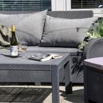 Außenbereich mit grauem Rattansofa, Tisch, Flasche und Buch.