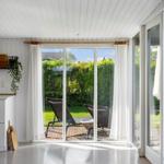Große Fensterfront mit Blick auf Garten und Liegestühle. Weiße Wände und Boden. Runder Teppich.