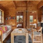 Wooden house with sofa, table, chairs, and wood stove. Windows and bookshelf visible.