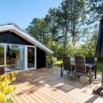 Deck with table, chairs, and closed umbrella. Background: forest and house with glass facade.