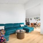 Ein Wohnzimmer mit türkisfarbenem Sofa, Holzboden und runden Couchtisch. Im Hintergrund ist ein Essbereich.
