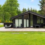 Modern black cabin with large wooden terrace, garden, and view of forest.