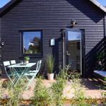 Black wooden cabin with terrace, table, chairs, and seating. Plants and garden area.
