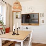 Dining room with wooden table, chairs, and wall-mounted TV. Window with plants and wall lamps.
