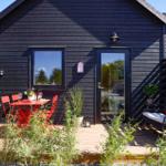 Black wooden house with terrace, red table and chairs, seating area and plants.