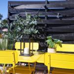 Yellow garden area with table, chairs, plants, and lantern in front of black wooden fence.