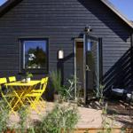 Black wooden house with terrace, yellow table and chairs, plants, and open entrance.