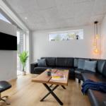 Ein Wohnzimmer mit schwarzem Sofa, Holztisch und Wand-TV. Große Fenster und helle Wände.