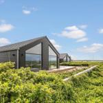 Moderne Holzhäuser mit großen Glasfronten in einer grünen Landschaft unter blauem Himmel.
