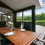 Terrasse mit Holztisch, Stühlen und Blumenarrangement. Blick auf grüne Hecke und gepflasterten Weg.