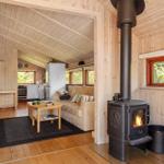 Living area with wood stove, sofa, and kitchen. Wooden walls and windows with view of greenery.