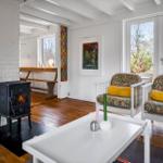 Living room with fireplace, two chairs, and hardwood floor. Windows overlook garden.