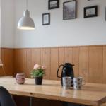 Wooden table with coffee mugs, pot, and flowers against wooden wall and window.
