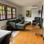 Wohnzimmer mit Sofa, Sessel, Tisch und Holzboden. Fenster mit Jalousien. Wandbild und Pflanzen.