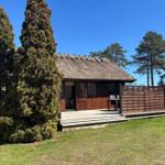Holzhütte mit Strohdach und Terrasse. Umgeben von Bäumen und Gras.
