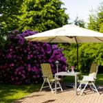 Terrasse mit Tisch, zwei Stühlen und Sonnenschirm. Hintergrund: grüne Bäume und lila Blüten.
