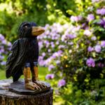 Holzfigur eines Vogels auf Baumstumpf im Garten mit lila Blumen.
