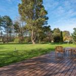 Wooden terrace with seating, surrounded by grass and trees under blue sky.