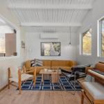A room with a sofa, piano, and windows. Wooden floor and white walls.