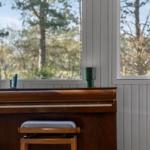 A piano with a stool in front of windows overlooking trees.