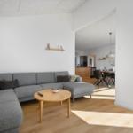 Ein Wohnzimmer mit grauem Sofa, runden Tisch und Holzboden. Durchgang zur Küche und Terrasse.