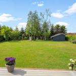 Large green garden with playground, shed, and flower beds. Wooden terrace in the foreground.