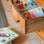 Holzschrank mit geöffneter Schublade, die Kinderbücher und Puzzles enthält, auf Holzboden mit Teppich.