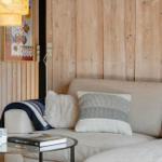 Ein Sofa mit Kissen und Decke vor einer Holzwand. Auf einem Tisch stehen Tassen und ein Buch.