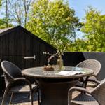 Terrasse mit runden Tisch und Stühlen, dunkler Holzwand und Bäumen im Hintergrund.