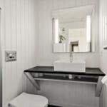 Badezimmer mit Waschbecken, Spiegel und Toilette. Weiße vertikale Wandverkleidung und schwarzem Unterschrank.