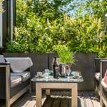 Terrasse mit Sitzgruppe, Tisch und Pflanzen. Hintergrund: grüne Hecke und Fenster.