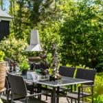 Außenterrasse mit Tisch, Stühlen und Grill, umgeben von grünen Pflanzen und Bäumen.