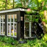 Ein kleines Haus mit großen Fenstern steht im Grünen. Eine Tür führt ins Innere.