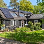 Schwarzes Holzhaus mit Solarpanelen, Terrasse und Garten. Umgeben von Bäumen und Gras.