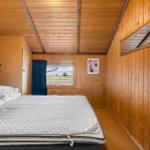 Schlafzimmer mit Holzwand, Bett und Fensterblick auf Landschaft.