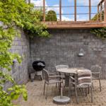 Gartenbereich mit Grill, Tisch und Stühlen. Hintergrund: Steinmauer und Holzterrasse.