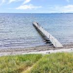 Langer Steg führt ins Meer. Menschen stehen dort. Strand und Gras sind sichtbar.